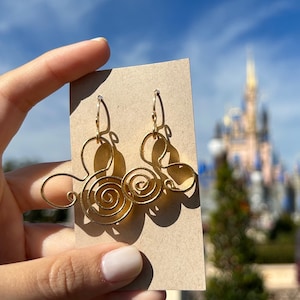 May include: Gold-colored wire earrings with a spiral design are held against a blue sky background. The earrings are on a tan card. The background includes a castle and trees.