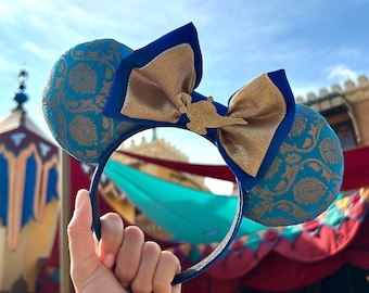 May include: Blue and gold Aladdin-themed mouse ears. The ears are a patterned teal fabric with a gold design. A gold bow with a gold lamp charm sits between the ears. The headband is blue.