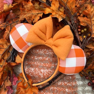 May include: Orange and white gingham mouse ear headband with a large orange bow. The headband is set against a backdrop of fall leaves and foliage, creating a seasonal theme. The ears are round and the bow is centered.