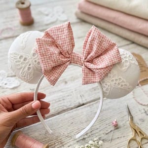 May include: A white headband with two round, white, lace-covered ears and a pink gingham bow. The headband is decorated with a pink gingham bow and white lace ears. The background includes sewing supplies and fabric.