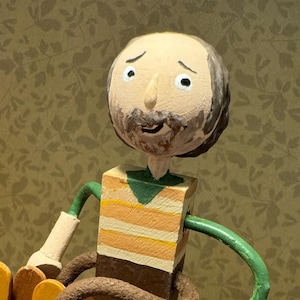 May include: A wooden figurine of a man with a beard, wearing a striped shirt, sitting on a fence with a pig standing on a stack of orange circles. The man is holding a green hose.