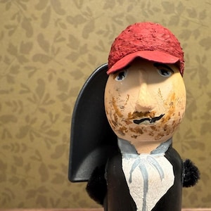 May include: A handcrafted wooden figurine of a man wearing a red cap, black coat, and blue pants. The figurine has a painted face with a mustache and is holding a black object. A glass jar with a lid is in the foreground.