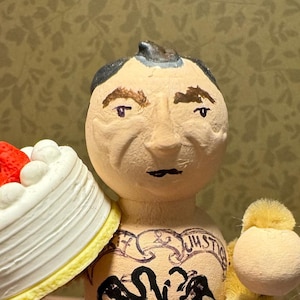 May include: A whimsical wooden figurine of a man holding a cake topped with strawberries and a basket of red cherries. The figurine has a tattoo that says "JUSTICE" and a small sign with the numbers "33379" and "390" and the date "D.C. 9-2-28".