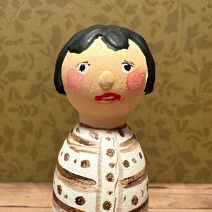 May include: A hand-painted wooden doll with black hair, a white and brown striped shirt, and a painted face with rosy cheeks.
