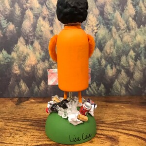 May include: A small orange figurine of a person with black hair, holding a pink and white checkered bag. The figurine is standing on a green base with a pile of trash, including a soda can, a plastic bottle, and some paper. The base is signed "Lisa Cain".