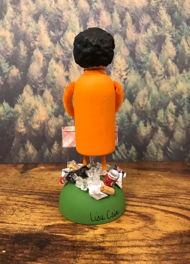 May include: A small orange figurine of a person with black hair, holding a pink and white checkered bag. The figurine is standing on a green base with a pile of trash, including a soda can, a plastic bottle, and some paper. The base is signed "Lisa Cain".