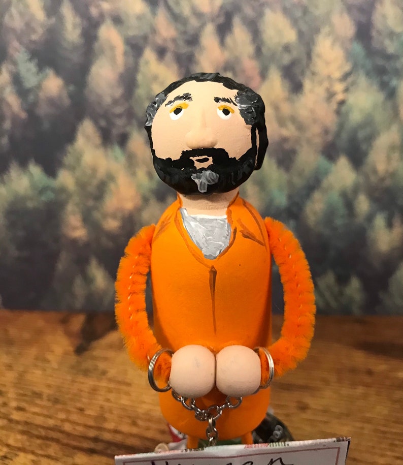 May include: Hand-painted figurine of a person wearing an orange jumpsuit and handcuffs. The figurine has a black beard and yellow eyes.