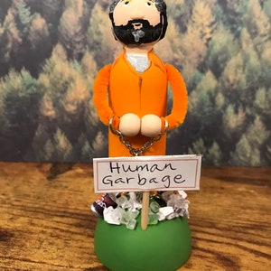 May include: A handmade doll figure of a person wearing an orange jumpsuit and handcuffs. The doll is standing on a green base with a sign that reads "Human Garbage". The base is surrounded by trash, including a can, plastic bags, and green and white crystals.