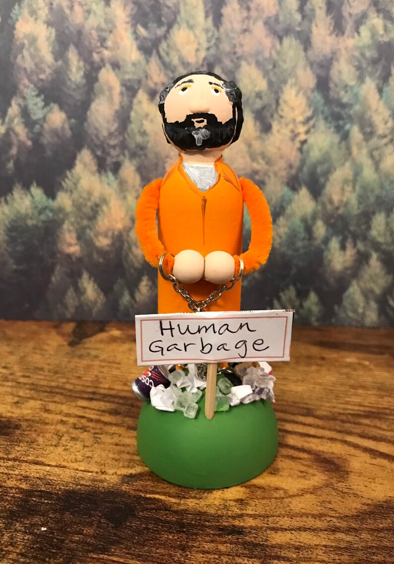 May include: A handmade doll figure of a person wearing an orange jumpsuit and handcuffs. The doll is standing on a green base with a sign that reads "Human Garbage". The base is surrounded by trash, including a can, plastic bags, and green and white crystals.