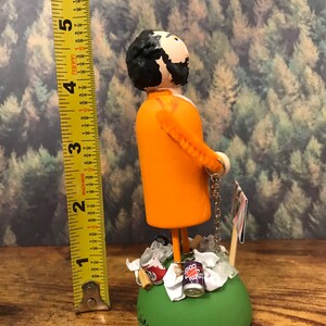 May include: A hand-painted figurine of a person wearing an orange jumpsuit and holding a chain. The figurine is standing on a green base with a variety of trash items around it, including cans, paper, and a wooden stick. The figurine is approximately 6 inches tall.