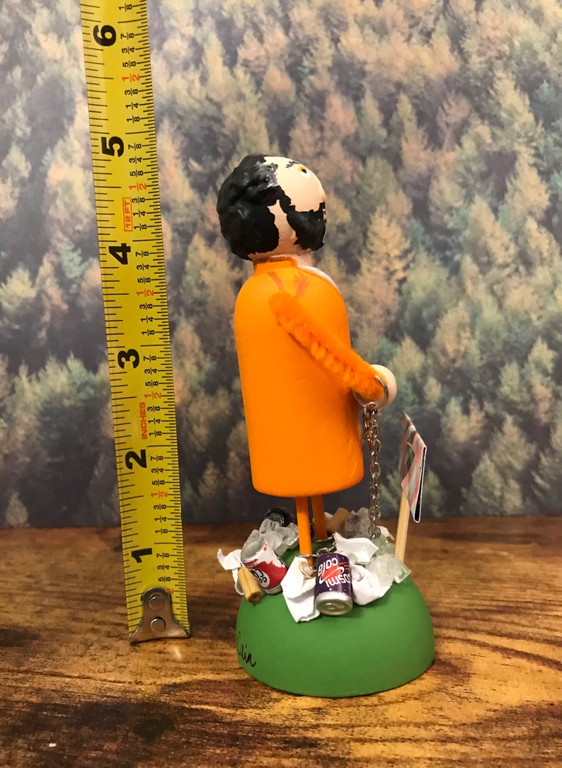 May include: A hand-painted figurine of a person wearing an orange jumpsuit and holding a chain. The figurine is standing on a green base with a variety of trash items around it, including cans, paper, and a wooden stick. The figurine is approximately 6 inches tall.