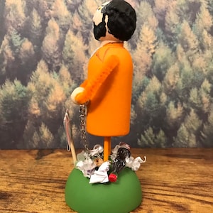 May include: A wooden figurine of a person wearing an orange jumpsuit and a chain around their ankle. The figurine is standing on a green base with a variety of small objects around it, including a soda bottle, a piece of paper, and a few white beads.