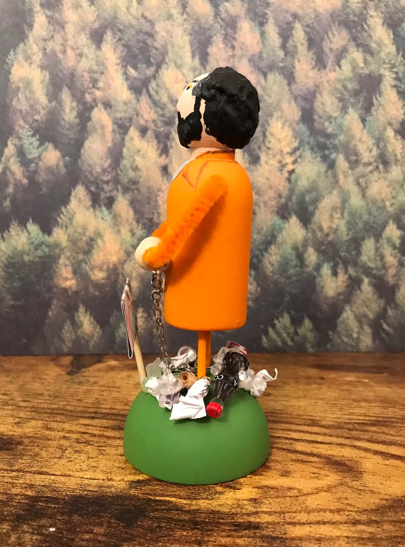 May include: A wooden figurine of a person wearing an orange jumpsuit and a chain around their ankle. The figurine is standing on a green base with a variety of small objects around it, including a soda bottle, a piece of paper, and a few white beads.