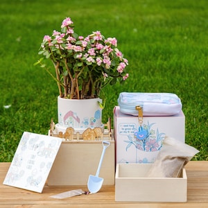 May include: A pink and white flower plant in a white ceramic pot with a cartoon house design. The plant is part of a gardening kit with a wooden box, a small blue shovel, and a card that says "Tiny Garden Card". The box has a blue and white zippered pouch with the text "I need help to grow all pretty till we grow old together".