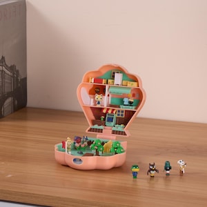 May include: A pink and green plastic playset shaped like a seashell, open to reveal a miniature house with furniture and characters. The playset is on a wooden surface with five small figurines.