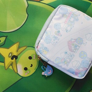 May include: A square, light blue and white patterned pouch with a zipper closure and a charm. The pouch is on a green and yellow patterned surface. The charm is blue and pink.