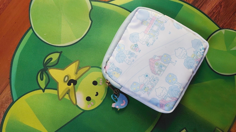 May include: A square, light blue and white patterned pouch with a zipper closure and a charm. The pouch is on a green and yellow patterned surface. The charm is blue and pink.