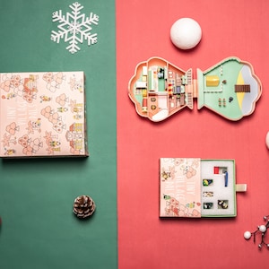 May include: A pink and green plastic dollhouse with a white snowflake ornament, a red ornament, a pine cone, and a box with a pink and white floral pattern. The dollhouse is open and shows the interior of the house, which is decorated with furniture and accessories.