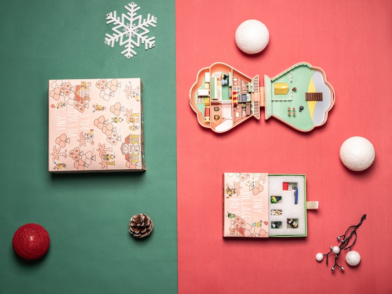 May include: A pink and green plastic dollhouse with a white snowflake ornament, a red ornament, a pine cone, and a box with a pink and white floral pattern. The dollhouse is open and shows the interior of the house, which is decorated with furniture and accessories.