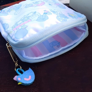 May include: A light blue zippered pouch with a cartoon design. The pouch has a gold-colored zipper pull and a felt bird charm. The interior features pink and blue stripes and a small cartoon character.