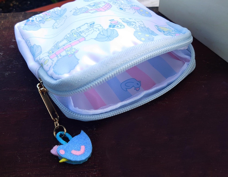 May include: A light blue zippered pouch with a cartoon design. The pouch has a gold-colored zipper pull and a felt bird charm. The interior features pink and blue stripes and a small cartoon character.