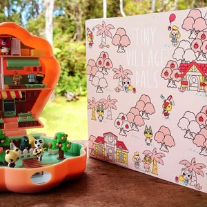 May include: A pink and white card with the text "Tiny Village Pals" and illustrations of animal characters from the Animal Crossing video game series.  The card is next to a small, orange plastic toy house with a hinged lid. The toy house is open and shows a miniature interior with furniture and animal characters.