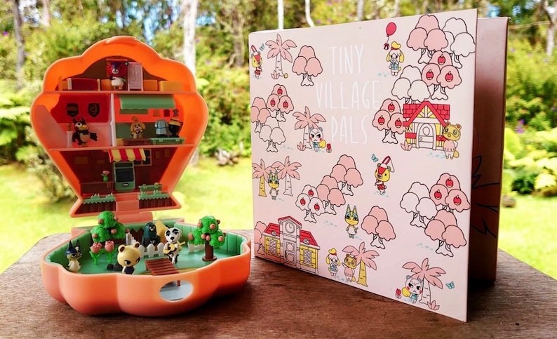 May include: A pink and white card with the text "Tiny Village Pals" and illustrations of animal characters from the Animal Crossing video game series.  The card is next to a small, orange plastic toy house with a hinged lid. The toy house is open and shows a miniature interior with furniture and animal characters.