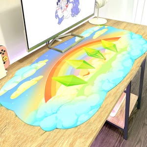 May include: A large, rectangular mouse mat with a colourful rainbow design, fluffy clouds, and green diamond shapes. The mat has a bright, cartoonish aesthetic, suitable for a gaming setup or workspace.
