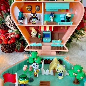 May include: A pink and blue plastic toy house with a green roof and a white picket fence. The house has a red flag, a brown wooden dock, and a yellow tent. There are several small plastic figures of animal characters from the Animal Crossing video game series.