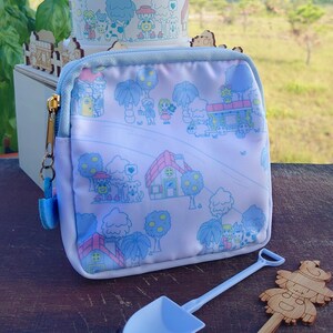 May include: A light blue zippered pouch with a whimsical village scene print, a matching ceramic planter, a small blue shovel, and a wooden decorative figure. The pouch has a light blue trim and a gold zipper.