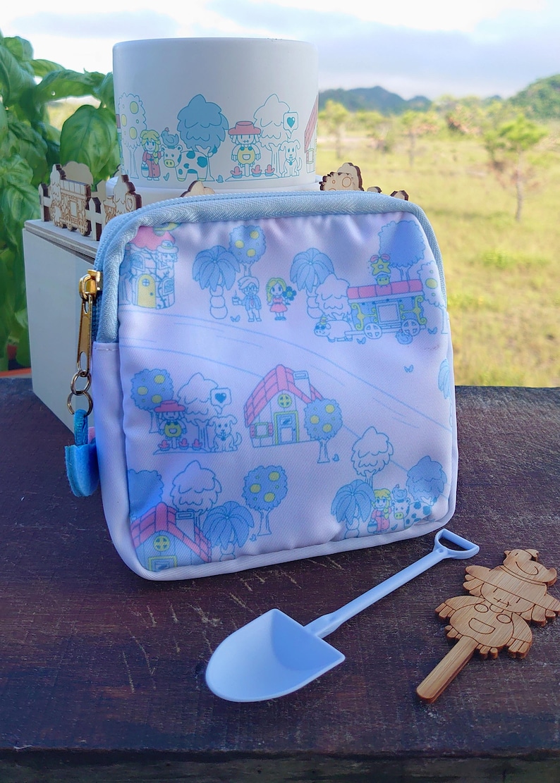 May include: A light blue zippered pouch with a whimsical village scene print, a matching ceramic planter, a small blue shovel, and a wooden decorative figure. The pouch has a light blue trim and a gold zipper.
