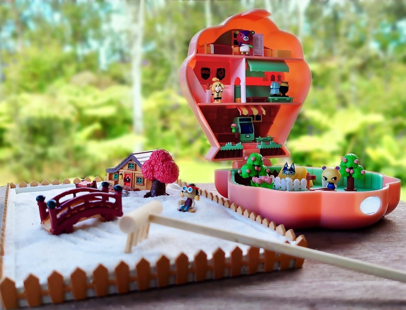 May include: A miniature dollhouse with a pink exterior and a white sand garden. The dollhouse is open and shows a multi-level interior with miniature furniture and figures. The garden features a red bridge, a rake, and a small tree.