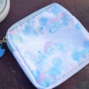 May include: A square, white pouch with a light blue zipper and trim. The pouch features a whimsical illustration in light blue and pink of a town with houses, trees, and a train. A blue bird charm hangs from the zipper.