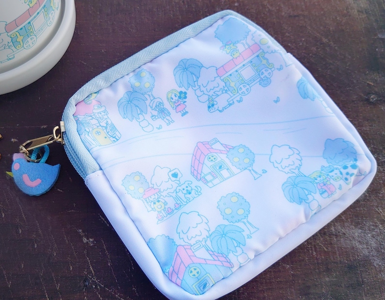 May include: A square, white pouch with a light blue zipper and trim. The pouch features a whimsical illustration in light blue and pink of a town with houses, trees, and a train. A blue bird charm hangs from the zipper.