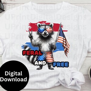 Puede incluir: Camiseta blanca con un mapache con gafas de sol con un dise&ntilde;o de la bandera estadounidense. El mapache sostiene una bandera estadounidense y lleva pantalones cortos rojos, blancos y azules. El texto de la camiseta dice "FERAL AND FREE".