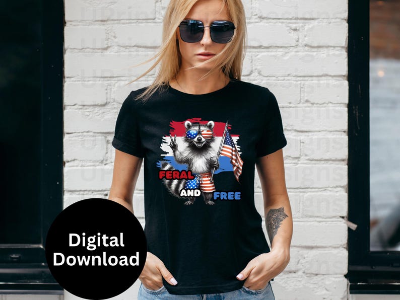 Puede incluir: Camiseta negra con un dise&ntilde;o de mapache con gafas de sol y ropa patri&oacute;tica, sosteniendo una bandera estadounidense. El texto "FERAL AND FREE" se muestra debajo. El dise&ntilde;o utiliza una combinaci&oacute;n de rojo, blanco y azul.