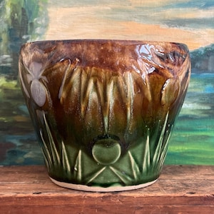 Può includere: Un vaso di fiori in ceramica vintage con finitura lucida. Il vaso presenta una ricca sfumatura di colore, che passa da un bordo marrone scuro a una base verde scuro. Sono visibili all'esterno motivi floreali e geometrici decorativi.