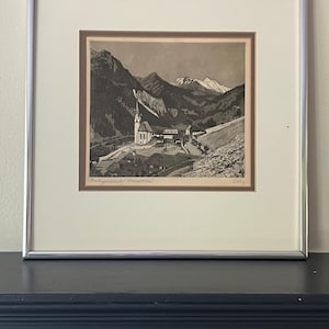 May include: A framed black and white etching depicting a mountain village with a prominent church, surrounded by steep slopes and snow-capped peaks. The artwork is set within a cream-coloured mat and a thin, polished silver-toned metal frame.