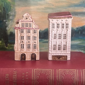 Op de afbeelding: Twee miniatuur keramische huisbeeldjes. Eén is een hoog, smal gebouw met een bruin dak. De andere is een breder gebouw met een decoratieve top. Beide zijn geschilderd in crème- en roze tinten, met gedetailleerde raam- en deurontwerpen.