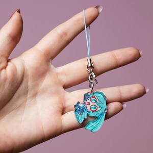 Könnte beinhalten: Ein kleiner Acryl-Anhänger mit einer stilisierten Anime-Figur mit türkisfarbenem Haar und großen Augen. Der Anhänger ist an einer hellblauen Kordelschlaufe mit einem silbernen Metallverschluss und einem kleinen silbernen Ring befestigt.
