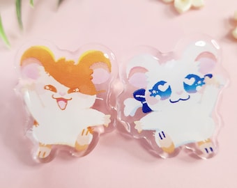 May include: Two clear acrylic pins featuring cartoon hamsters. One hamster is white with blue accents, the other is orange and white. Both have smiling faces and are waving. The background is a soft pink.