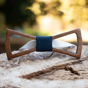 Puede incluir: Una pajarita de madera con un centro de tela azul oscuro y una banda de tela blanca. La pajarita está sentada sobre un trozo de madera desgastada.