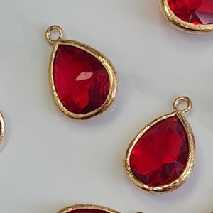 May include: Red teardrop-shaped pendants with gold-colored metal edging. The pendants are faceted, giving them a sparkling appearance. Each pendant has a small loop at the top for attaching to jewelry. The pendants are scattered across a light-colored surface.