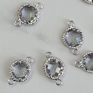 Puede incluir: Una colección de conectores de vidrio gris facetado con marcos de metal plateado. Cada conector tiene un centro de vidrio redondo y transparente con un borde de metal retorcido decorativo y pequeños bucles para sujetar. Los conectores están dispersos sobre una superficie blanca.