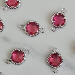 May include: Scattered across a light surface are multiple silver-toned connectors with a round, faceted, magenta-colored gem. The gems are set in a twisted rope-like border. Each connector has a small loop on either side.