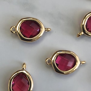 Puede incluir: Primer plano de varios componentes de joyería dorados con piedras preciosas rojo rubí ovaladas y facetadas. Cada componente tiene un lazo a cada lado para la fijación. Las piedras preciosas están engastadas en un bisel dorado.