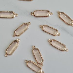 May include: Close-up of several rectangular gold-toned metal charms with a light pink, translucent center. Each charm has a loop on each end for attaching to other jewelry components. The charms are scattered on a light-colored surface.