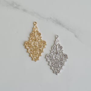 Op de afbeelding: Twee sierlijke filigraanhangers, één goudkleurig en één zilverkleurig, worden getoond tegen een lichte achtergrond. Elke hanger heeft een gedetailleerd, symmetrisch ontwerp met een lus aan de bovenkant om op te hangen. Geschikt voor het maken van sieraden.