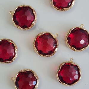 May include: Circular, faceted, ruby-red gemstone pendants with gold-coloured, textured bezels. Each pendant has a small loop at the top for attaching to jewellery. The pendants are scattered across a light-coloured surface.