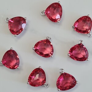 May include: Eight triangular, faceted, ruby-red gemstone pendants are displayed. Each pendant features a silver-coloured bail and three small silver-coloured prongs. The gemstones are arranged on a light-coloured surface.